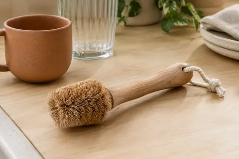 Brosse tawashi à tête conique en fibre de coco, manche en bois et boucle en coton posée près d’une tasse
