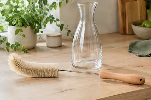Brosse longue en sisal et bois de hêtre posée près d’une carafe en verre sur un plan de travail clair